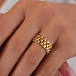 Puede incluir: Un anillo dorado con un diseño rectangular en forma de rejilla. El anillo está hecho de pequeños bloques cuadrados dispuestos en un patrón repetitivo. El anillo está en un dedo.