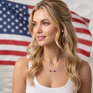 Pode incluir: Um colar de tom dourado com um pingente de barra horizontal apresentando três pedras retangulares em vermelho, branco e azul. O colar é usado com uma regata branca e brincos, com uma bandeira americana desfocada ao fundo.