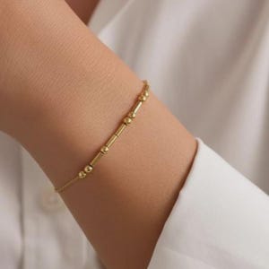 Peut inclure: Un bracelet doré délicat composé d'une série de petites perles rondes et de barres cylindriques. Le bracelet est porté sur un poignet, avec une chemise blanche en arrière-plan. Le bijou est simple et élégant.