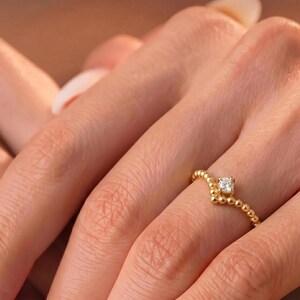May include: A gold ring featuring a delicate beaded band with a V-shaped center setting. A single small, clear, round-cut gemstone is mounted at the peak of the V-shape, creating a subtle focal point on the thin, textured metallic band.