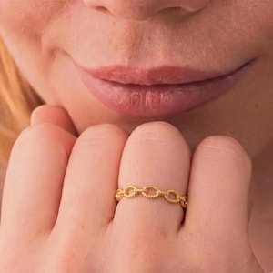 May include: A close-up of a gold ring with a chain link design. The ring is worn on a finger, with the person's lips and part of their face visible in the background. The ring is made of a shiny, yellow metal.