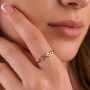 Può includere: Un anello in oro caratterizzato da un delicato design a fascia intrecciata a forma di infinito. L'anello in metallo è incastonato con piccole pietre chiare a taglio rotondo inserite a intervalli regolari lungo il centro della struttura in filo d'oro.