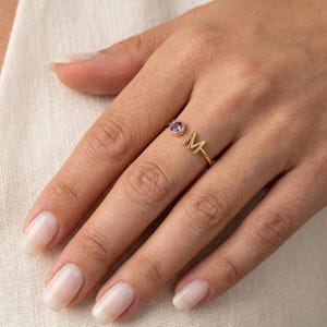 Op de afbeelding: Een goudkleurige ring met de letter "M" en een kleine paarse edelsteen. De ring wordt aan een vinger gedragen en toont zijn delicate ontwerp en elegante eenvoud. De edelsteen is rond en gezet in een bezel.