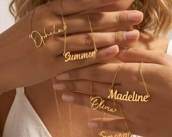 May include: Gold-colored name necklaces spelling out "Ophelia", "Summer", "Olivia", "Madeline", and "Simona". The necklaces are displayed against a soft background, showcasing the delicate script of each name.