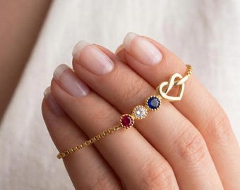 May include: A gold-colored bracelet featuring a heart-shaped clasp and a series of four colorful gemstones: red, clear, blue, and navy. The bracelet is designed with a delicate chain and is displayed on a person's hand.