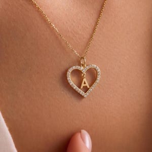 Pode incluir: Um colar dourado com um pingente em forma de coração. O pingente é adornado com pequenas pedras transparentes e apresenta a letra "A" no centro. A corrente e o pingente são dourados.