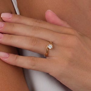 May include: A gold ring with a single, round, clear gemstone. The ring's band curves around the gemstone, creating a unique and elegant design. The ring is shown on a person's finger.