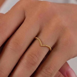 May include: A gold-colored ring with a V-shaped design. The ring is adorned with small, evenly spaced beads, creating a textured appearance. The ring is worn on a finger.