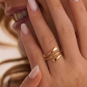 May include: A gold-colored ring with a spiral design. The ring wraps around the finger with two parallel bands and rectangular accents. The ring is worn on a hand with manicured nails.