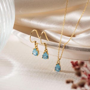May include: A gold-toned jewelry set consisting of a necklace and a pair of drop earrings. Each piece features a single light blue, pear-shaped gemstone pendant suspended from a delicate gold chain or a curved gold ear wire hook.