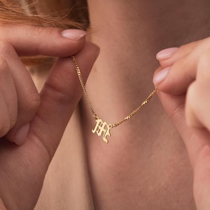 Op de afbeelding: Een goudkleurige ketting met een hanger in de vorm van de letter "M" in een oud-Engels lettertype. De ketting wordt door een persoon omhoog gehouden. De ketting is delicaat en de hanger is klein.