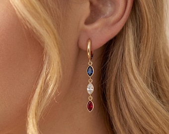 May include: Gold hoop earring with three dangling marquise-cut gemstones in blue, white, and red. The earring is shown on a person's ear, with the gemstones hanging below the earlobe. The earring is a stylish accessory.