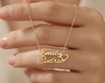 May include: A gold-colored necklace with the names "Emily" and "Cedric" in a cursive font, connected by a heart symbol. The necklace is held up by a person's hand, showcasing the delicate chain and pendant design.