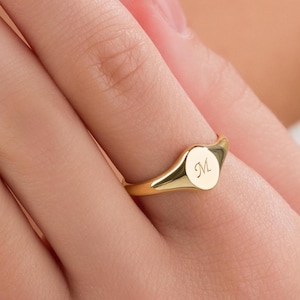 May include: A gold signet ring with a polished finish. The ring features a round face with the engraved letter "M". The ring is shown on a finger.