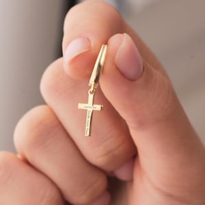 May include: A gold-colored cross pendant with the words "psalm 34" and "Faith over Fear" engraved on it. The cross is suspended from a curved, gold-colored earring hook. The jewelry is held up by a person's hand.