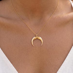 May include: A gold-colored crescent moon pendant necklace. The pendant has a white inner layer and hangs from a delicate gold chain. The necklace is worn against a light skin tone.