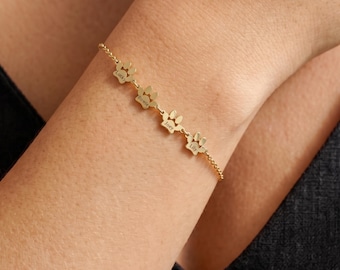 May include: A gold-toned chain bracelet featuring four connected paw print charms. Each charm is engraved with a name: Lucky, Bowie, Joey, and Lily. The delicate metal chain links the charms together in a linear sequence, creating a personalized piece of jewelry.