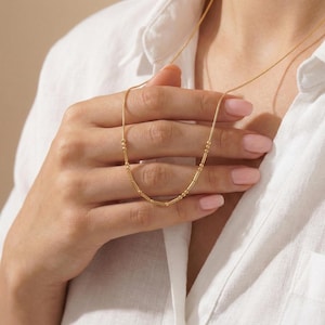 May include: A delicate gold necklace with small beads and bar-shaped elements. The necklace is shown against a white shirt, emphasising its simple elegance. The necklace is a stylish accessory.
