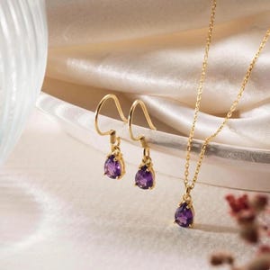 Può includere: Un set di gioielli in oro composto da un paio di orecchini pendenti e una collana con ciondolo abbinata. Ogni pezzo presenta una pietra preziosa di ametista viola a forma di goccia sospesa a una sottile catena d'oro e ganci d'oro lucidi.