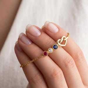 May include: A gold-tone chain bracelet featuring three round bezel-set stones in red, clear, and blue, followed by a small open heart-shaped charm. The delicate box chain design connects these decorative elements in a linear arrangement.