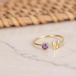 Pode incluir: Um anel dourado com uma pedra preciosa roxa e a letra "M". O anel está sobre uma superfície de mármore branco. O design é minimalista e elegante, adequado para o uso diário.