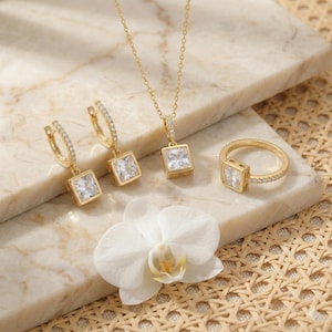 May include: A gold-toned jewelry set featuring a necklace, earrings, and ring, each with a square, clear gemstone. The earrings and ring are accented with small, sparkling stones. A white orchid flower rests near the jewelry.