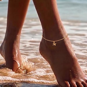 Op de afbeelding: Een goudkleurige enkelband met een fijne ketting en een bedel met de letter "M". De enkelband wordt gedragen op een enkel bij de oceaan. Het water is lichtblauw.