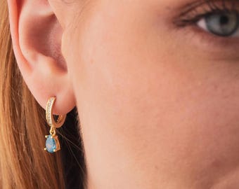 May include: A gold hoop earring featuring a row of small clear stones along the band and a single teardrop-shaped light blue stone dangling from the bottom. The hoop is secured through the earlobe, showcasing a polished metallic finish.