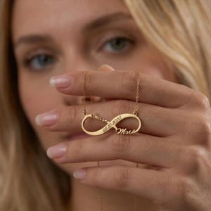May include: A gold-colored infinity necklace with the names "Susan" and "Mare" engraved on it, along with the date "05.10.2022". The necklace is held up by a person.