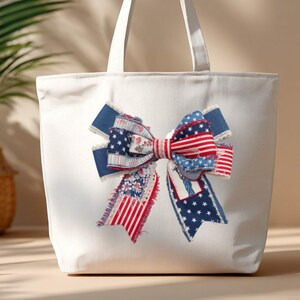 May include: A white canvas tote bag with a large bow applique. The bow features red, white, and blue fabric with stars and stripes, and floral patterns. The bag has two white handles.