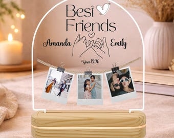 May include: A clear arched acrylic plaque with a wooden base featuring the text Best Friends, the names Amanda and Emily, a pinky promise illustration, and the text Since 1996. Three small photos are clipped to a string across the plaque.