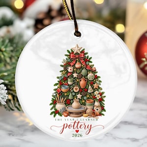 May include: A round white ceramic ornament featuring a Christmas tree decorated with miniature pottery, mugs, and tools. The text reads THE YEAR I LEARNED pottery 2026 with a small red heart. A black ribbon is attached to the top for hanging.