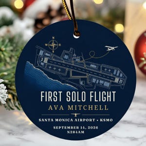 May include: A round dark blue ornament featuring a white line drawing of an airport runway map, a compass rose, and a small aeroplane. Text reads: FIRST SOLO FLIGHT, AVA MITCHELL, SANTA MONICA AIRPORT KSMO, SEPTEMBER 14, 2026, and N284AM.