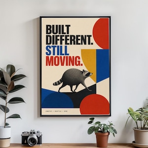 May include: A framed poster featuring a gray raccoon walking across a geometric background of red, blue, and yellow shapes. The text reads BUILT DIFFERENT. STILL MOVING. with JIMOTHY / SEATTLE / 2026 printed in small black letters at the bottom.