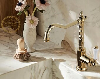 May include: A gold-colored faucet with a curved spout and white handle. A small wooden brush with bristles sits on the counter. A vase with pink flowers is in the background. The faucet has a classic design.