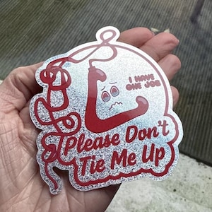 May include: A glittery sticker featuring a red, cartoon rope design with a face, and the text "I Have One Job" and "Please Don't Tie Me Up". The sticker is held in a hand.