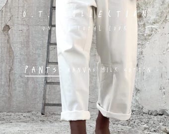 May include: Off-white canvas pants with rolled cuffs, part of a total look collection. The image includes text that reads "Pants: Canvas Milk Cotton". The model is wearing a white top. The background is a textured, light-colored wall.