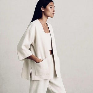 May include: Cream-colored linen jacket with a square neckline and large front pockets. The jacket is part of a matching set, paired with a crop top and high-waisted pants. The outfit is a neutral, minimalist style.