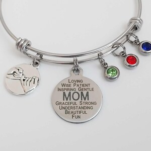 May include: Silver bangle bracelet with several charms. One charm shows a hand gesture, another has the word "MOM" and descriptive words. Additional charms include green, red, and blue gemstones.