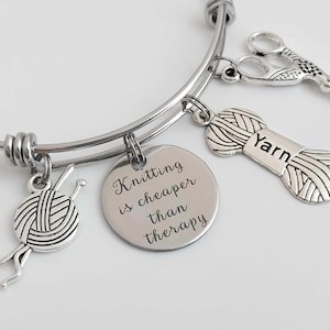 May include: Silver bangle bracelet with knitting-themed charms. Charms include a yarn ball with needles, a yarn spool with "Yarn" text, scissors, and a circular charm with the text "Knitting is cheaper than therapy."