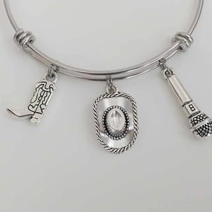 May include: Silver-toned bangle bracelet with three charms: a cowboy boot, a cowboy hat, and a microphone. The charms are suspended from a curved, adjustable band. The bracelet is designed to be worn on the wrist.