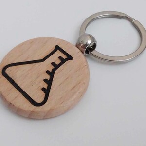 May include: A wooden keychain with a black outline of a beaker on a light brown background. The keychain is attached to a silver metal ring.
