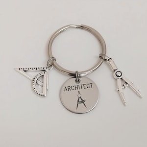 May include: Silver architect keychain with a round tag that reads "ARCHITECT" and a compass graphic. The keychain also features a small protractor and a compass charm. The keychain is on a silver ring.