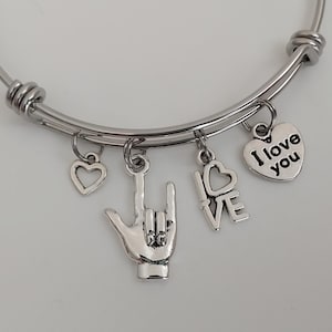May include: A silver bangle bracelet with multiple charms, including a heart, the sign for 'I love you' in American Sign Language, and a heart charm with the words 'I love you'.