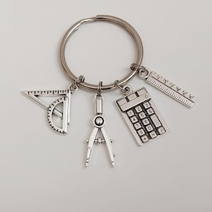 May include: Silver-toned keychain featuring charms of a protractor, compass, calculator, and ruler. The charms are attached to a circular key ring. A unique gift for students, engineers, or anyone who appreciates tools.