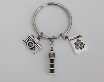 May include: Silver keychain with three charms: a camera, a miniature Big Ben tower, and a passport with the word "PASSPORT" printed on it.
