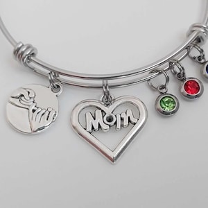 May include: Silver bangle bracelet with various charms. Charms include a heart-shaped pendant with the word "Mom", a round charm with hands, and three round charms with coloured gemstones: green, red, and blue.