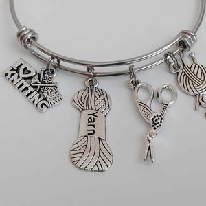 Könnte beinhalten: Ein silberfarbenes Armband mit Anhängern zum Thema Stricken. Zu den Anhängern gehören eine Stricknadel und ein Wollknäuel, eine Schere und ein Anhänger mit der Aufschrift "I Love Knitting". Der Anhänger mit der Aufschrift "yarn" ist ebenfalls enthalten.