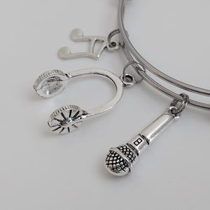 Op de afbeelding: Zilverkleurige bedelarmband met muziekthema. De armband heeft bedels in de vorm van een G-sleutel, een koptelefoon en een microfoon. De bedels zijn bevestigd aan een zilverkleurige, rekbare armband.