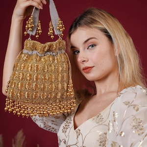 May include: A gold beaded drawstring bag with a grey handle and gold beaded accents. The bag has a detailed pattern and a fringe of gold beads. The background is a deep red.